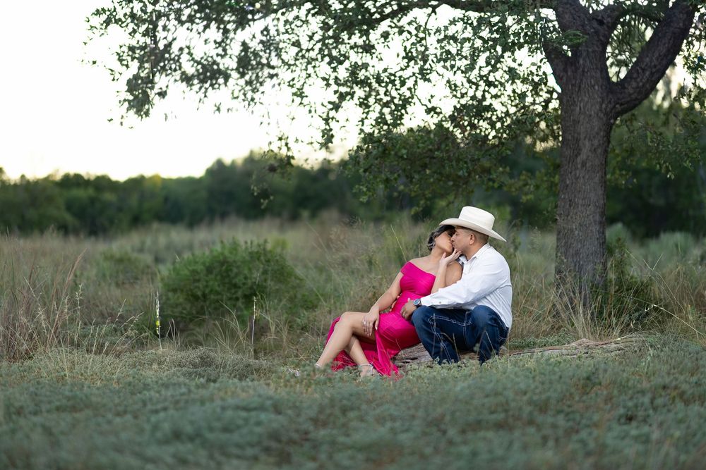 A couple relaxes together under a large tree on a grassy hill during sunset.