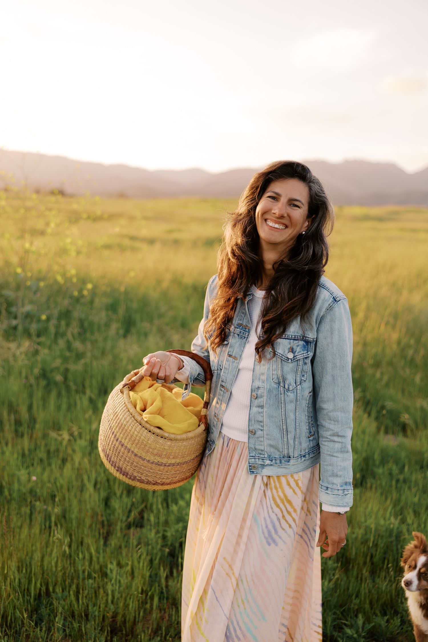 A person in a denim jacket and flowy dress stands in a sunny meadow holding a woven basket during golden hour.