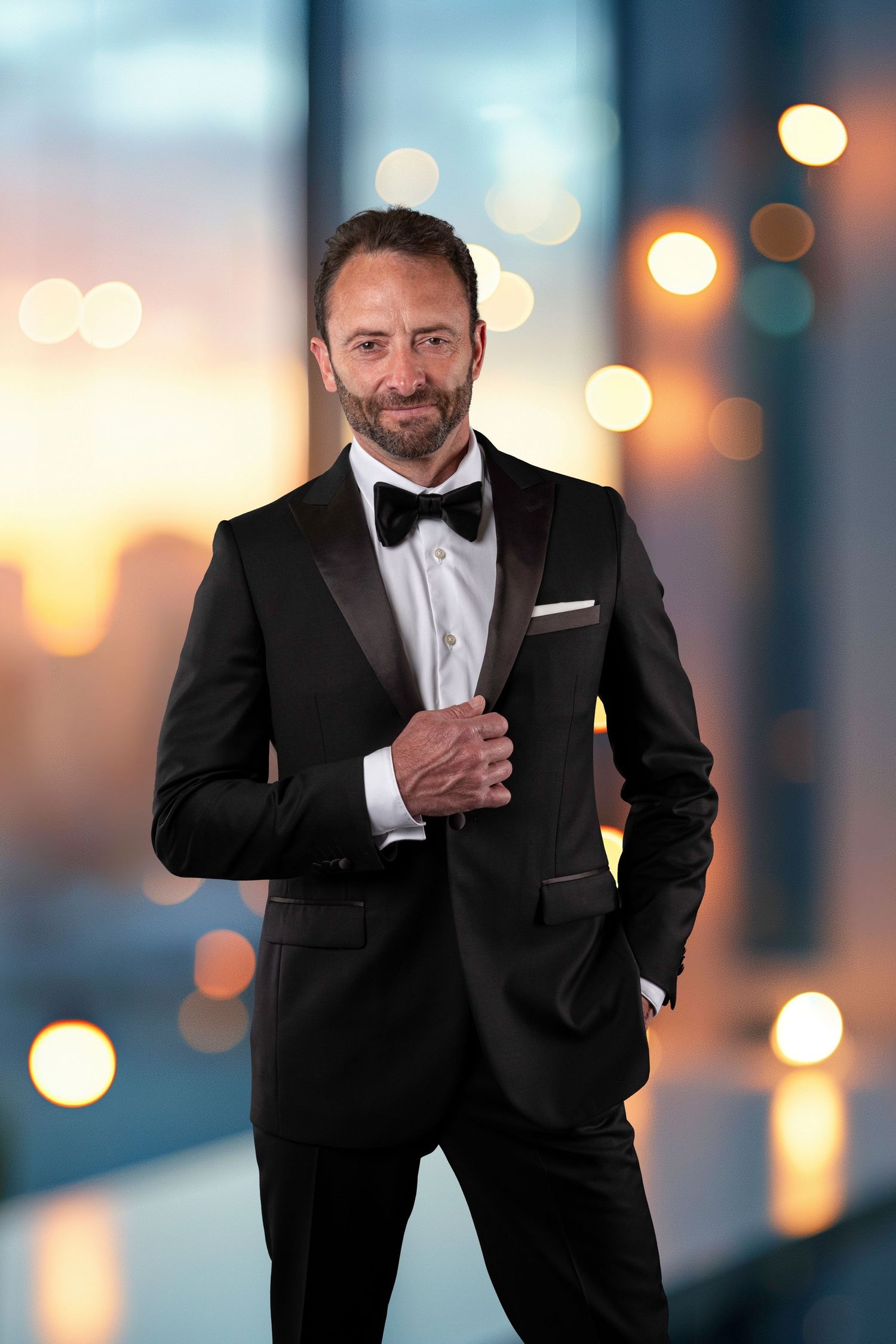 A person wearing a black tuxedo and bow tie poses confidently against blurred lights in a formal portrait setting.