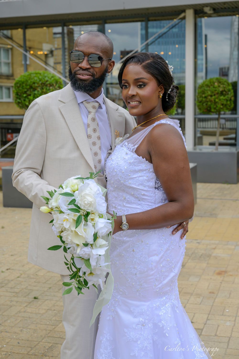 A wedding couple in a cream suit and white beaded dress pose with white lily bouquet on a stone paved courtyard.