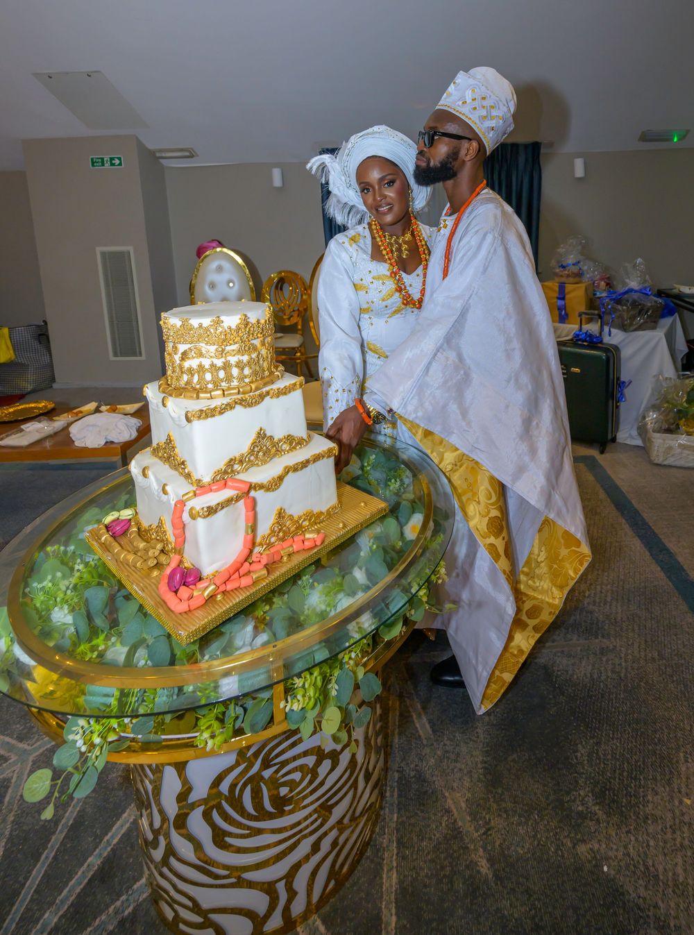 A couple in white traditional attire standing next to an ornate gold and white wedding cake.