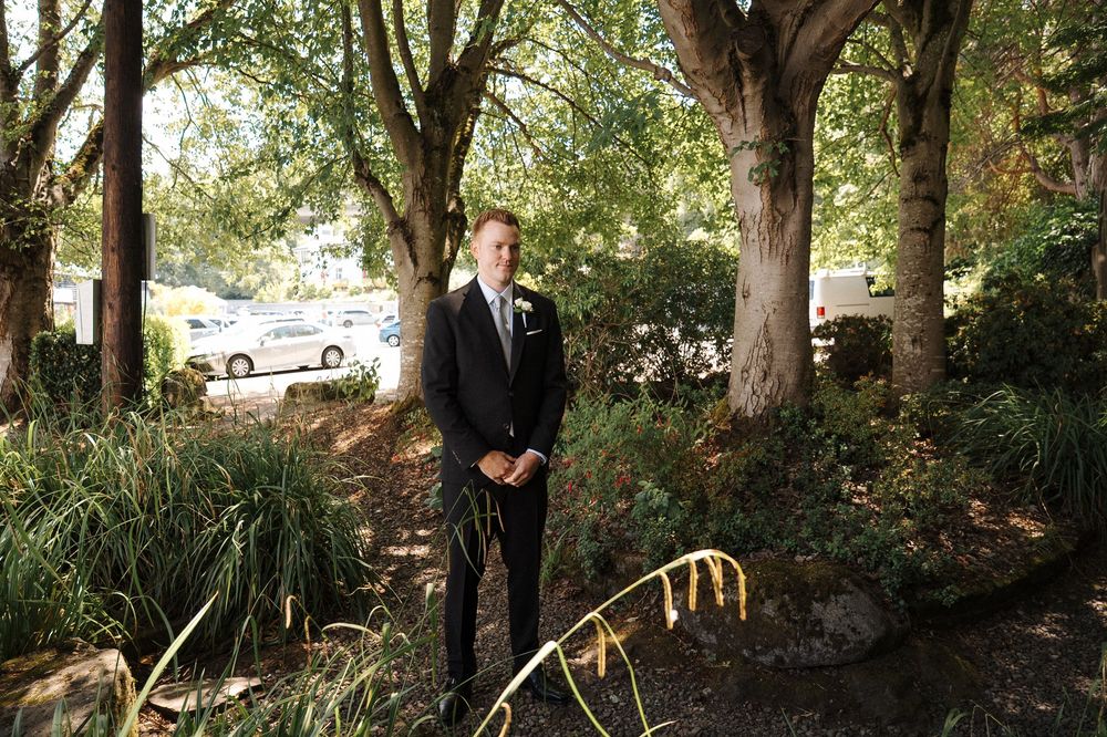 Elegantly dressed person stands in a tranquil garden setting surrounded by mature trees and natural landscaping.
