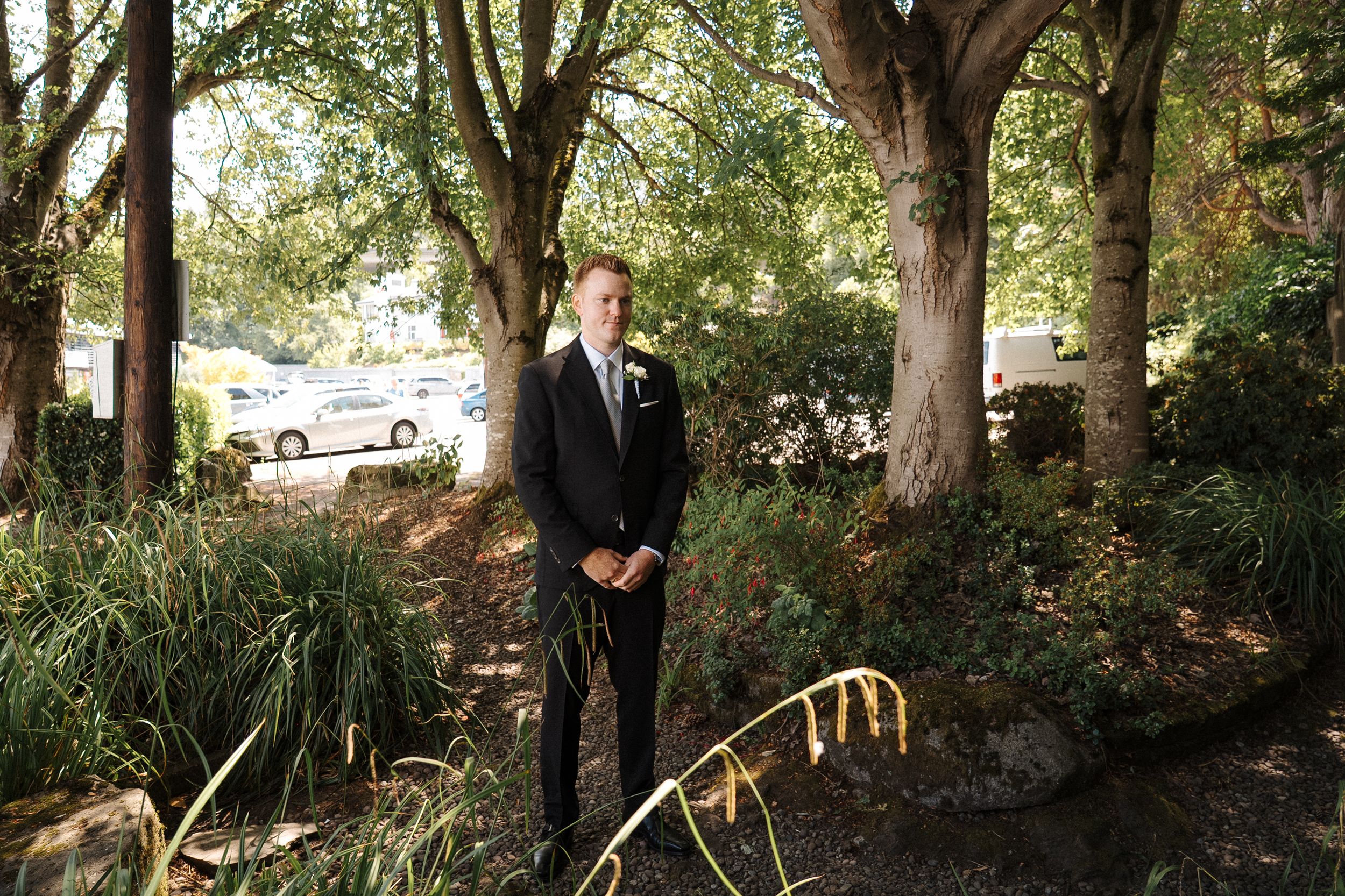 Elegantly dressed person stands in a tranquil garden setting surrounded by mature trees and natural landscaping.