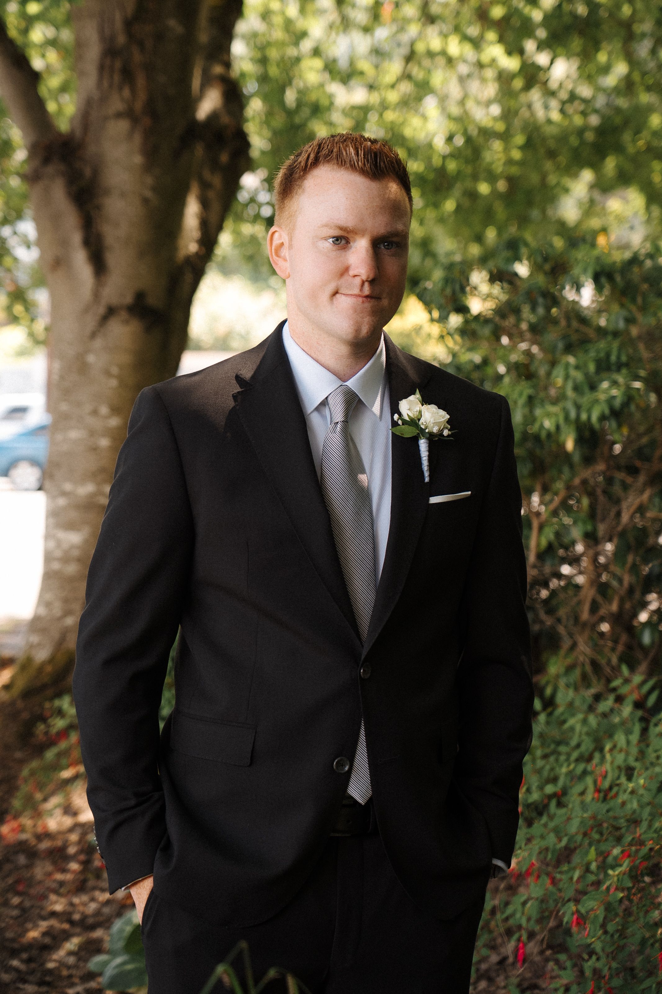 Wedding party members in black suits and white ties stand together under green trees in an outdoor setting.