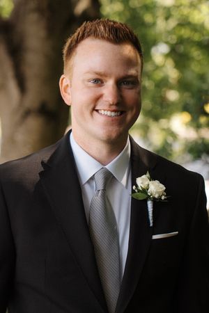 A groom in a black suit and gray tie smiling in an outdoor wedding portrait series with natural lighting.