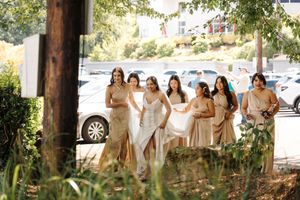 Wedding party gathers outdoors under leafy trees while posing for photographs in coordinated neutral attire.