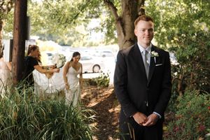 Elegant summer wedding scene in a garden setting with people in formal attire standing under shaded trees.