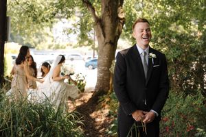 Elegant summer wedding scene in a garden setting with people in formal attire standing under shaded trees.