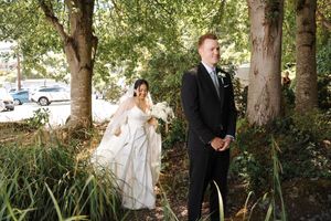 Outdoor wedding ceremony setup with guests seated under tall shady trees on a sunny afternoon.