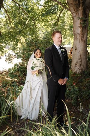 Newlywed couple shares candid moments together among trees and greenery in an outdoor garden setting.