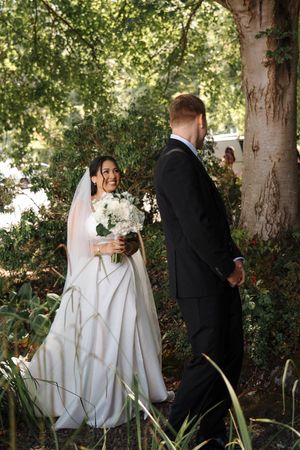 A bride in a white gown and groom in black suit share a first look moment in a romantic garden setting.