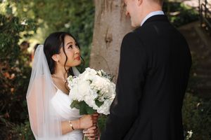 Two people in traditional wedding attire share a joyful moment outdoors holding a white floral bouquet.