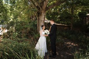 Couple in elegant wedding attire embrace beneath towering trees in a romantic outdoor forest setting.