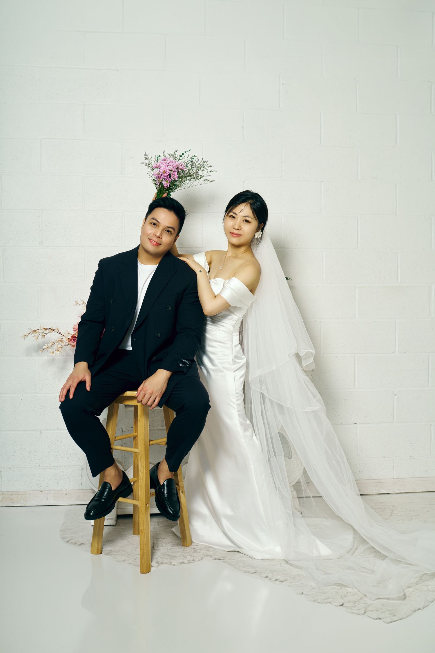 A wedding portrait against a white wall featuring a couple with a groom seated on a stool and bride in flowing white dress.