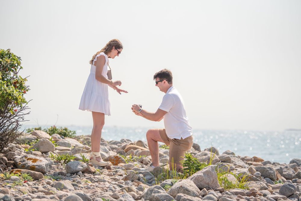 Surprise proposal scene on rocky beach with ocean in background.