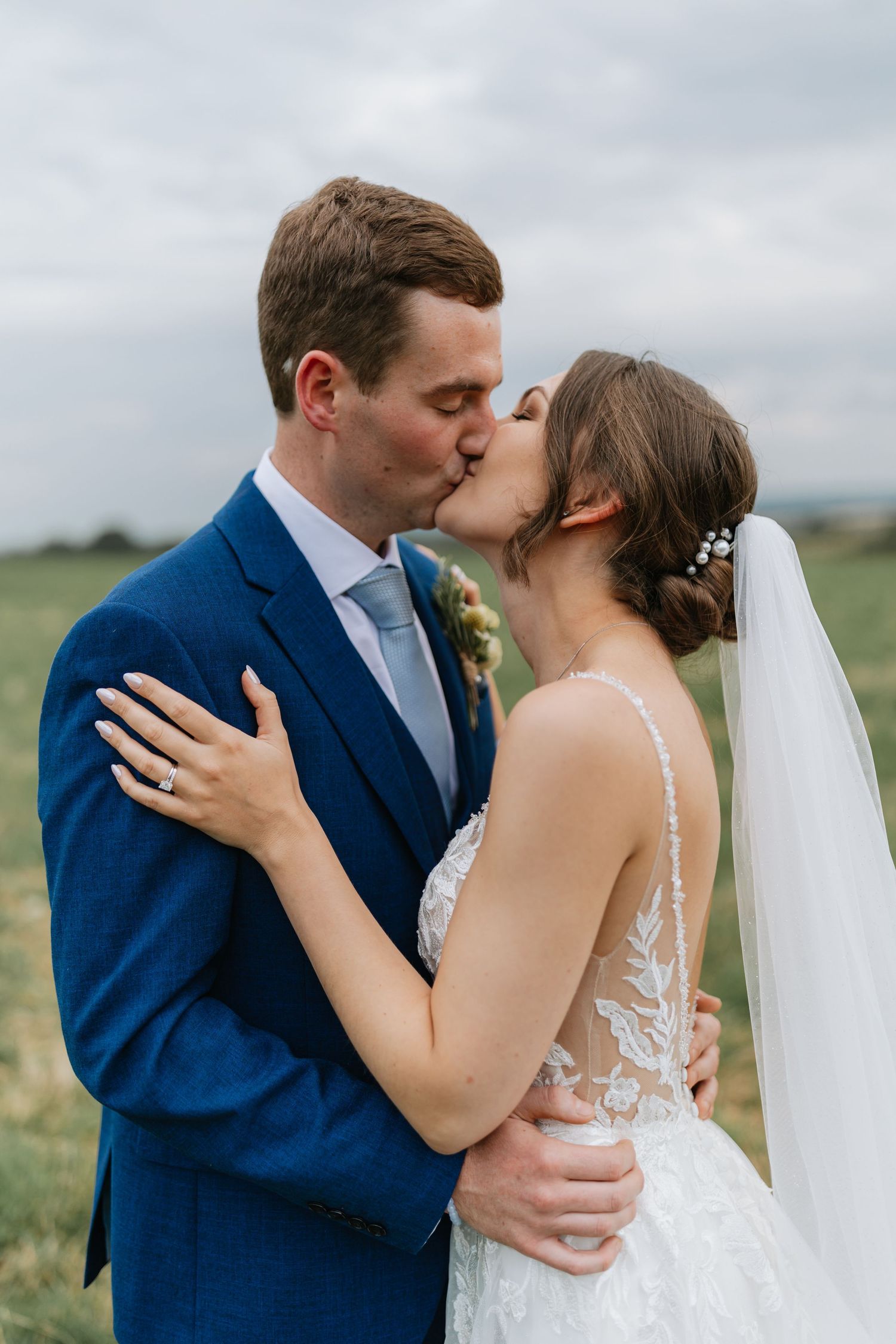 A romantic wedding moment showing a couple sharing a kiss outdoors with the bride in lace and groom in navy blue suit.