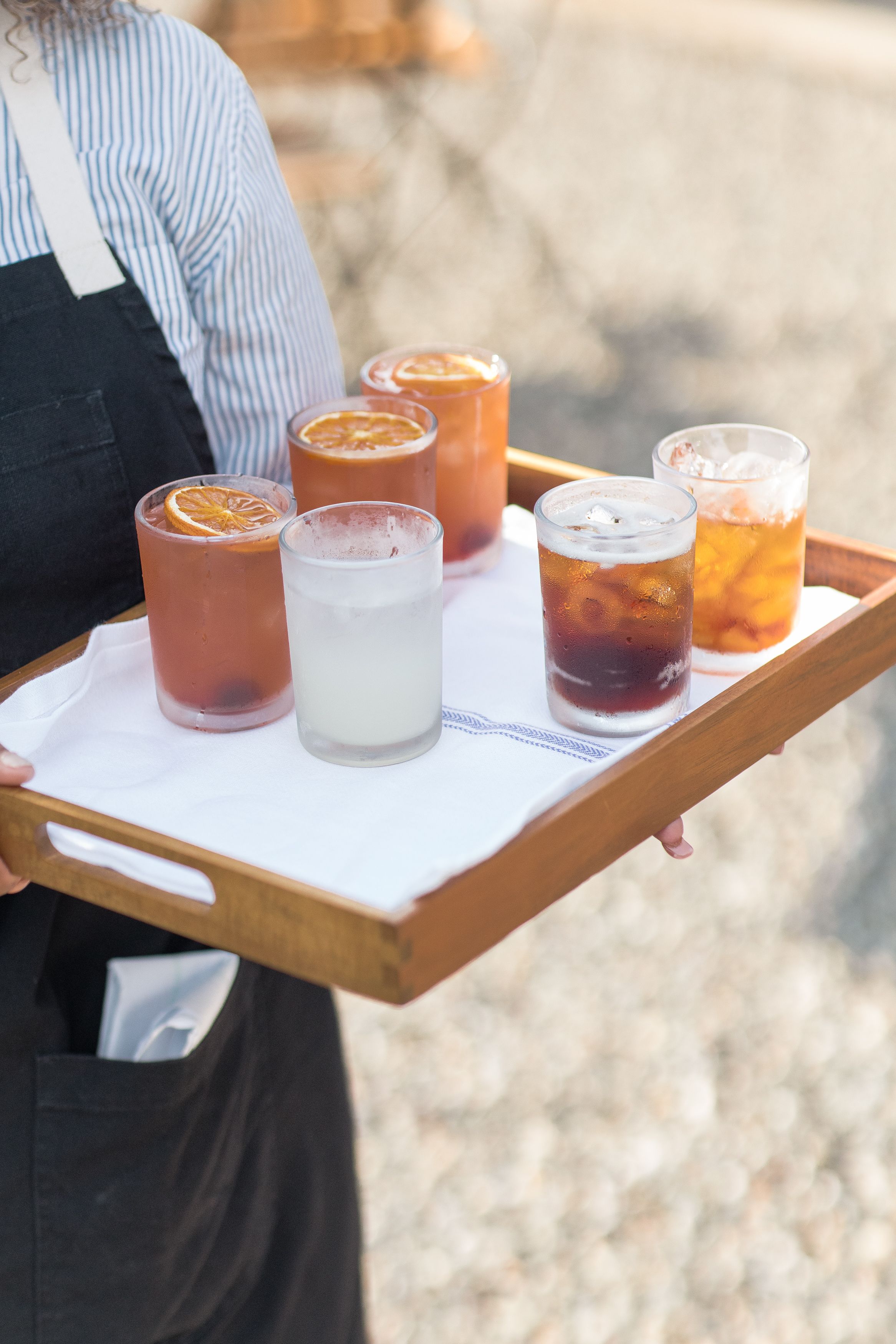 Server holding wooden tray with various colorful cocktails and beverages in small glasses at outdoor event.