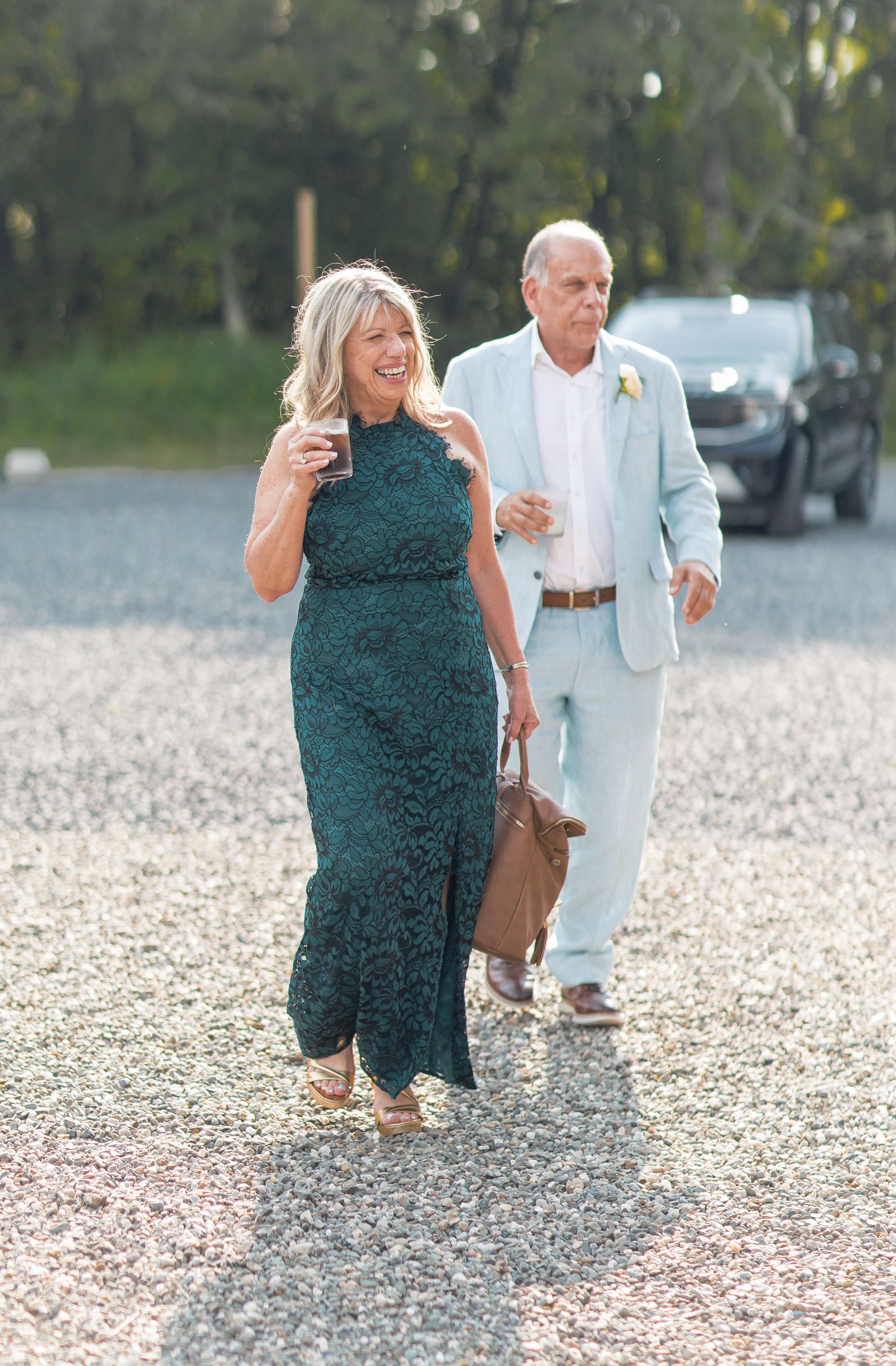Wedding guests walk on a pebbly beach path toward an outdoor celebration in elegant formal attire.