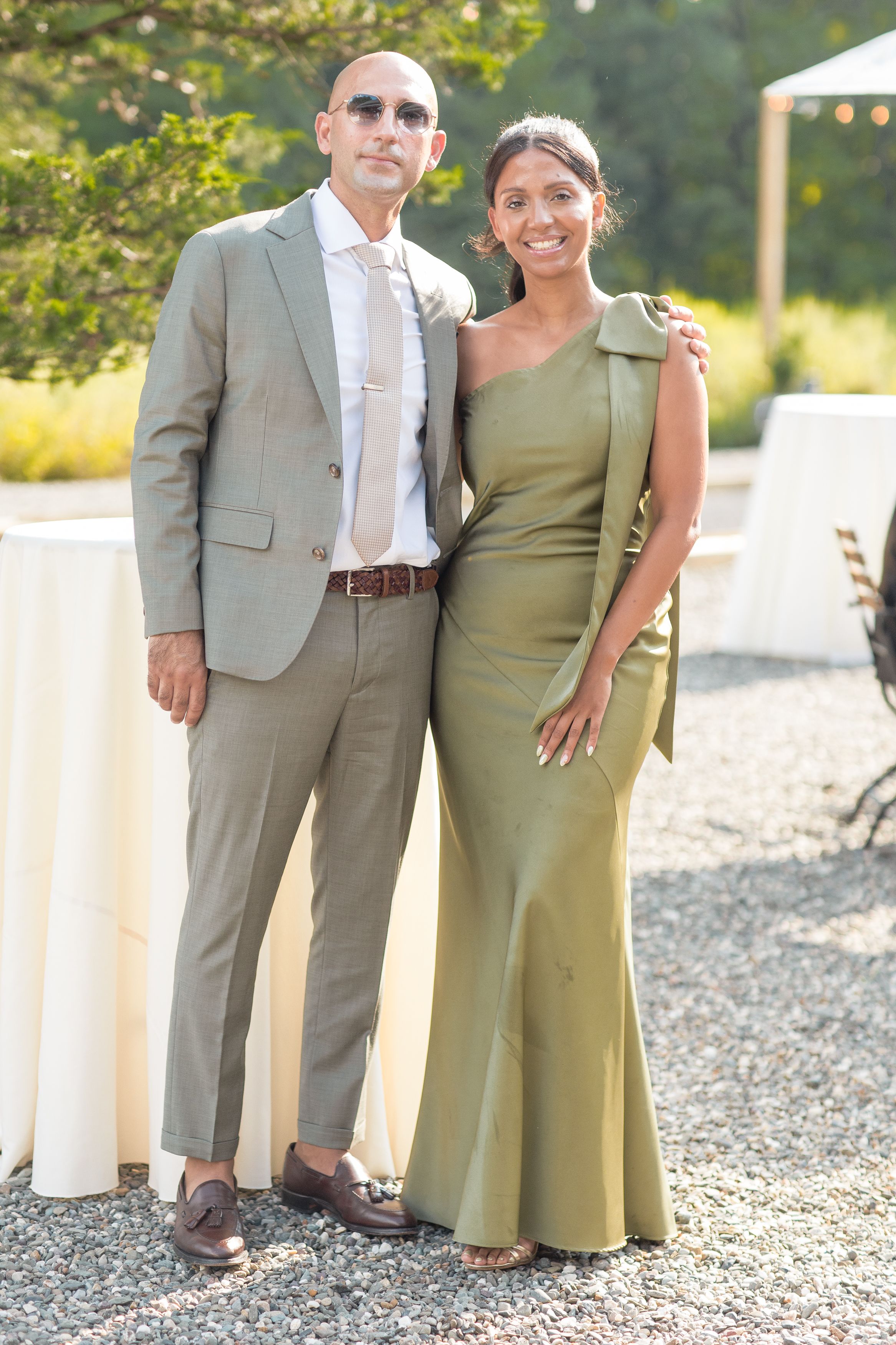 A couple poses casually outdoors on a gravel surface with sunlit trees in the background.