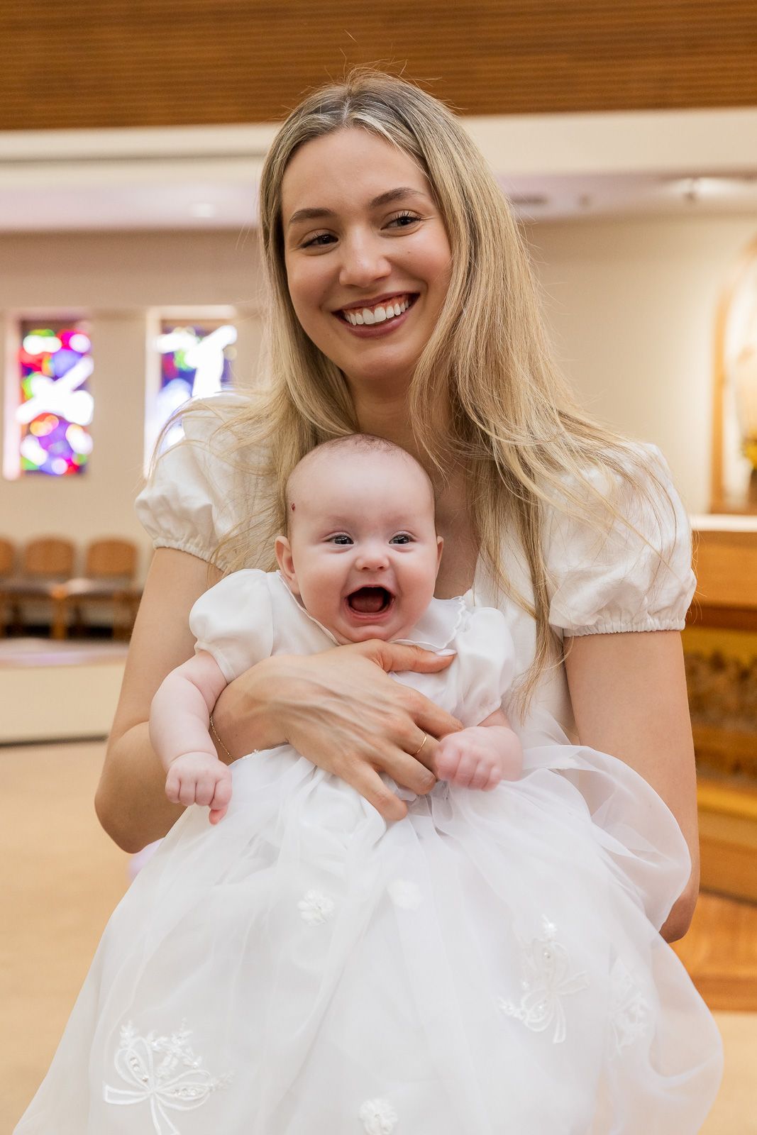 An adult and baby wearing matching white formal dresses share a joyful moment together during what appears to be a christening.