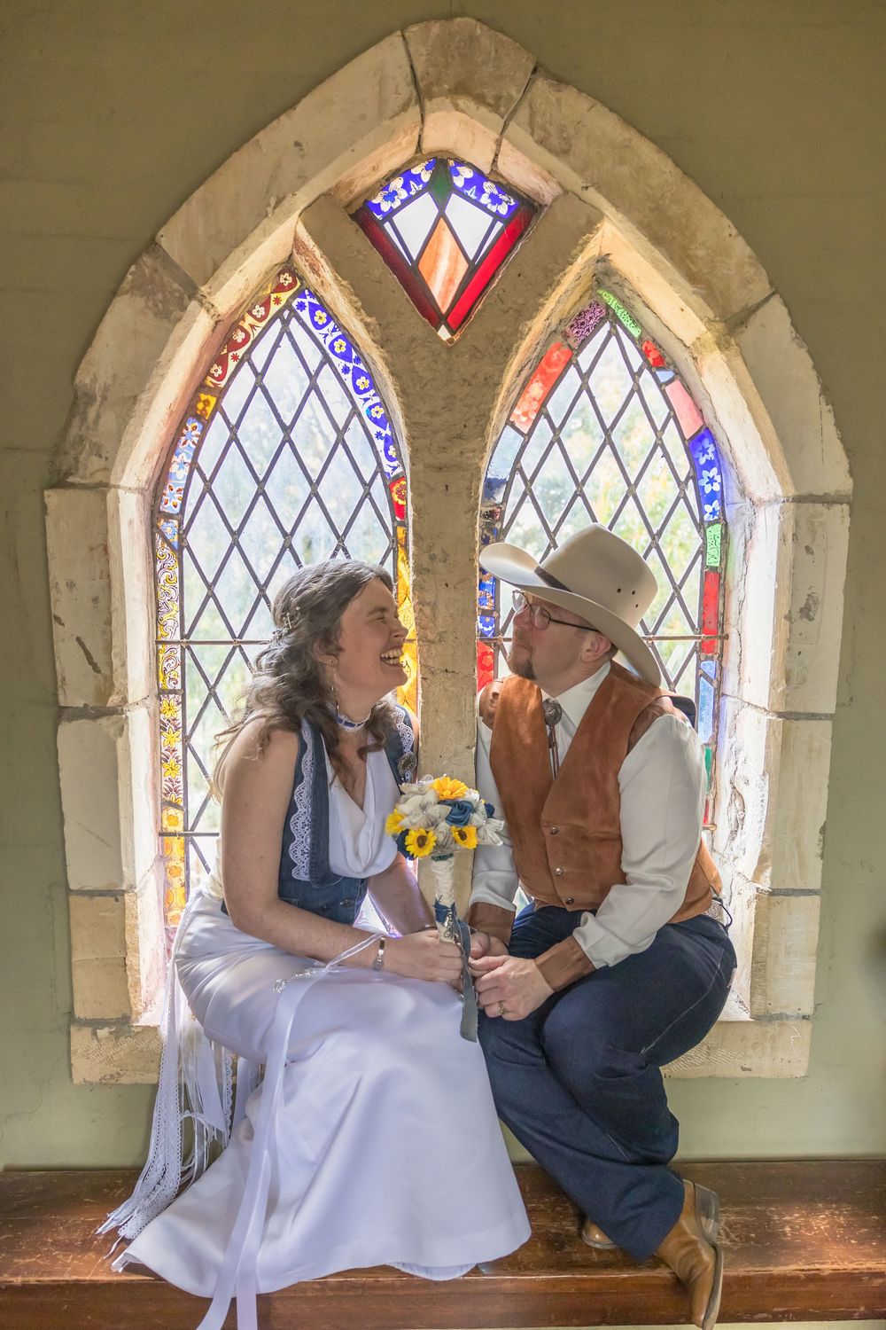 A couple shares a romantic moment while sitting in an arched church window with stained glass, surrounded by soft light.