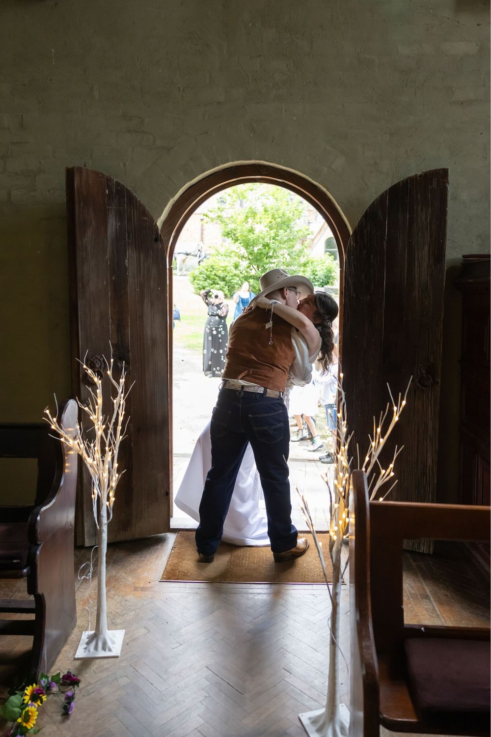 Wedding couple shares a romantic kiss in old rustic doorway of historic stone building with sunlight streaming through window.