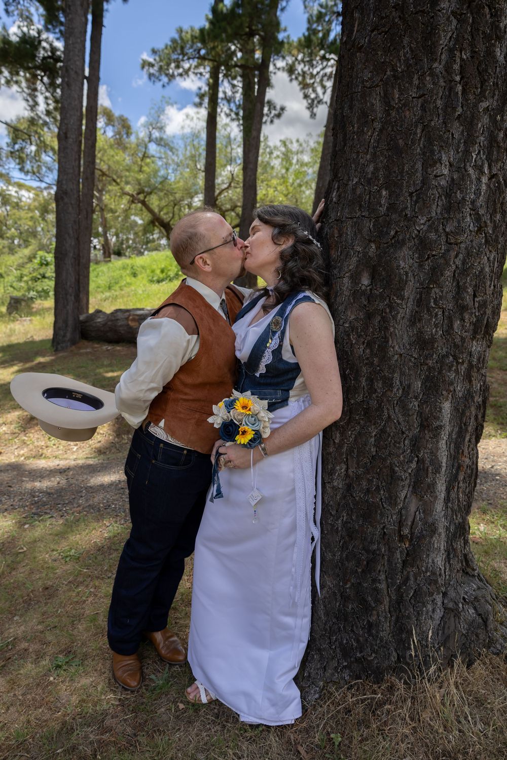 A romantic couple in western and wedding attire sharing a kiss in a wooded outdoor setting with pine trees.