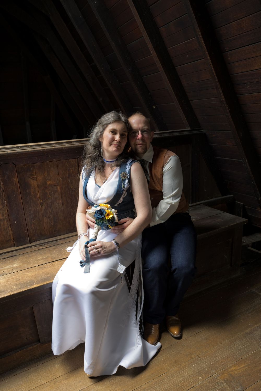 A couple in formal attire sits together on a wooden bench in a rustic attic space.