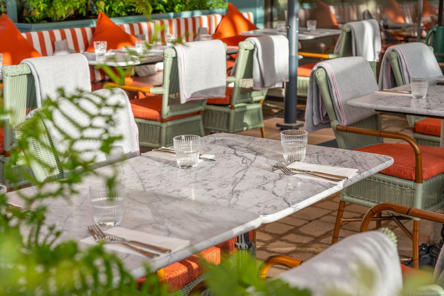 Outdoor restaurant dining area with orange cushions and white tablecloths set against lush greenery and brick walls.