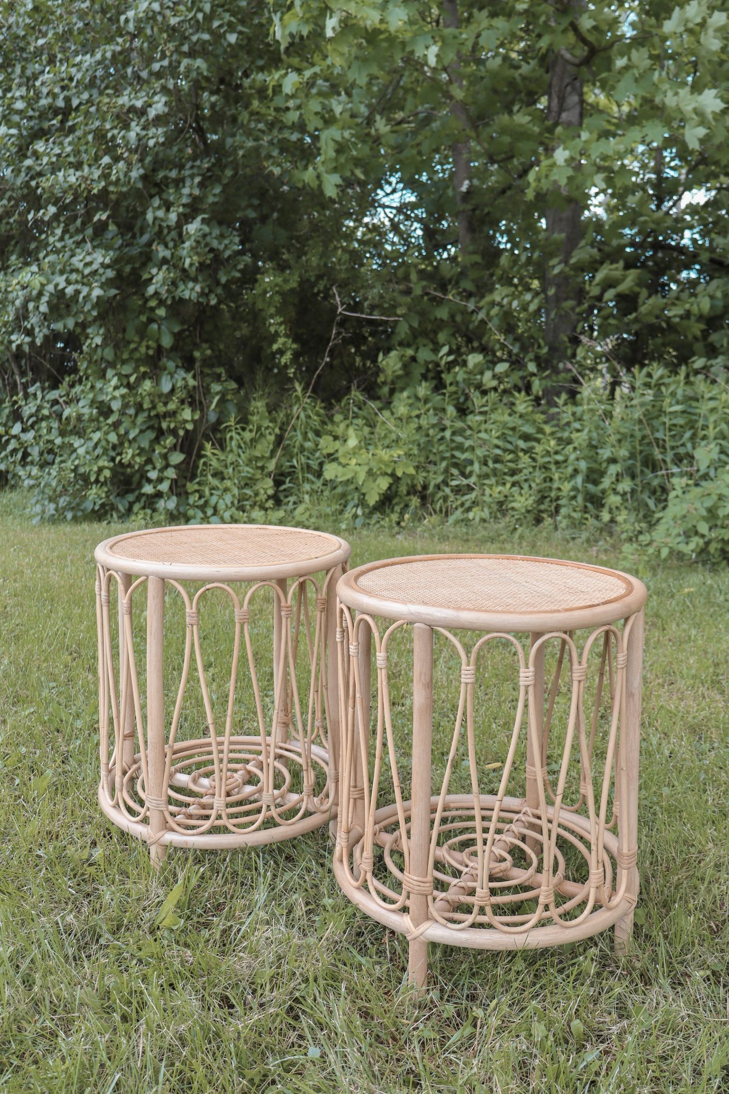 Two beige rattan side tables with round tops and curved architectural designs sit on green grass in an outdoor setting.