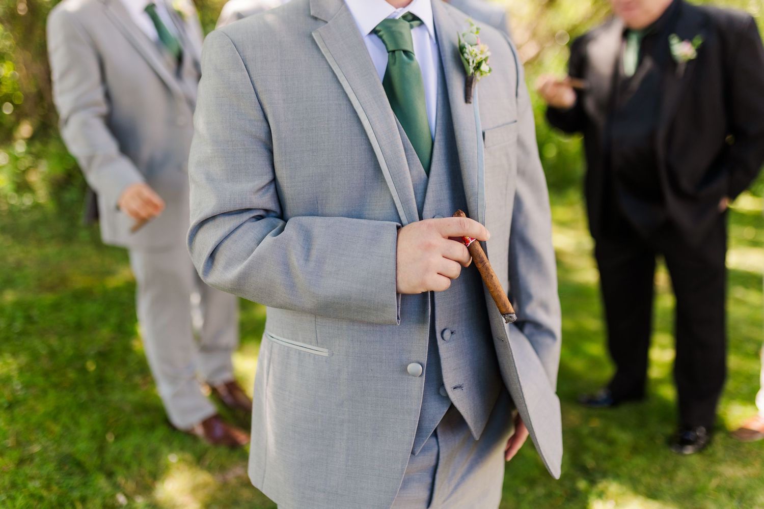 Groomsmen in light gray suits with green ties pose together on a grassy lawn during an outdoor wedding.