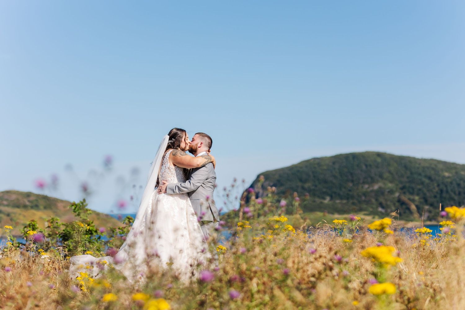A couple embraces in a wildflower meadow with mountains in the background on a sunny day.
