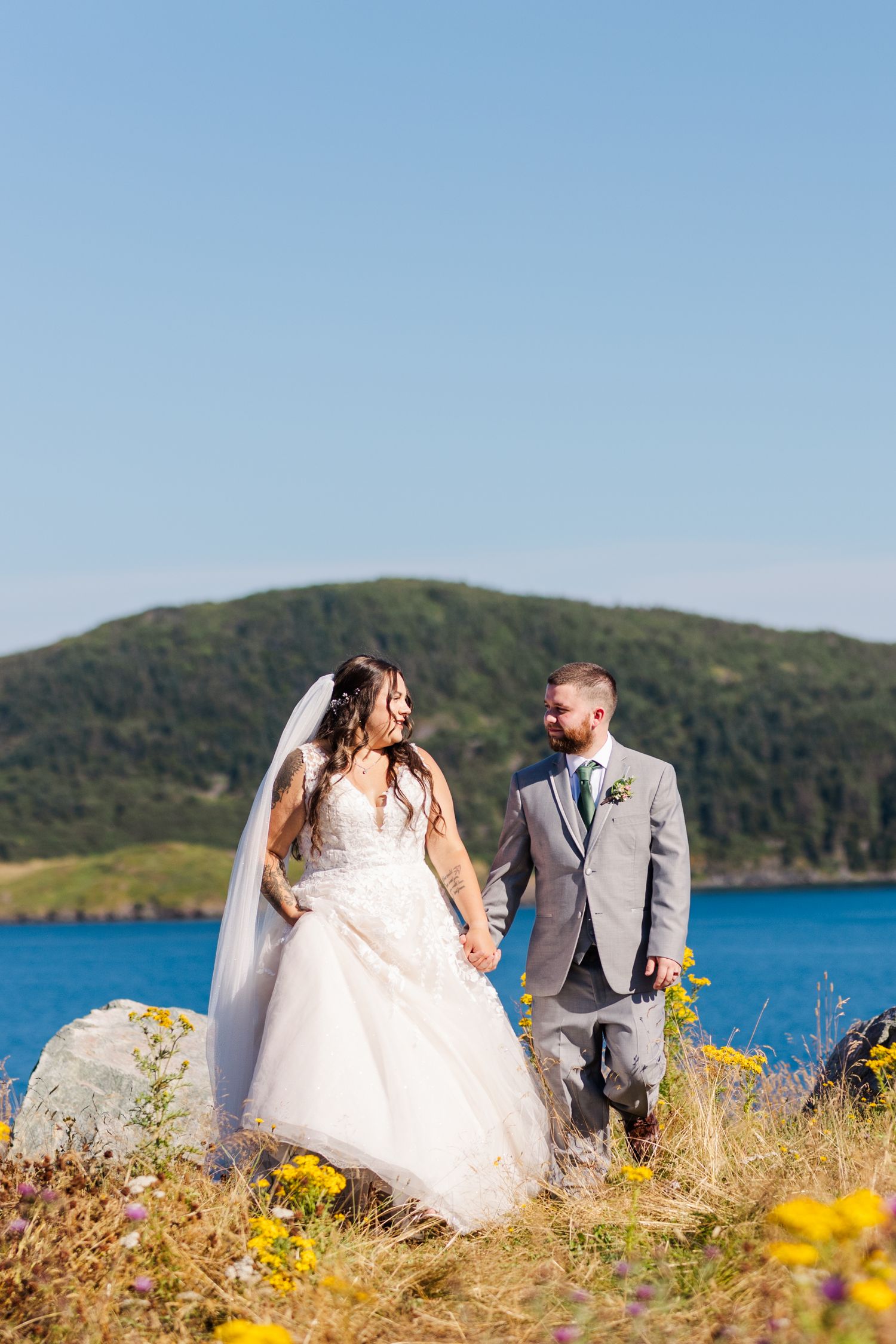 Couple in wedding attire standing on coastal hillside with blue water and mountains in background on sunny day.