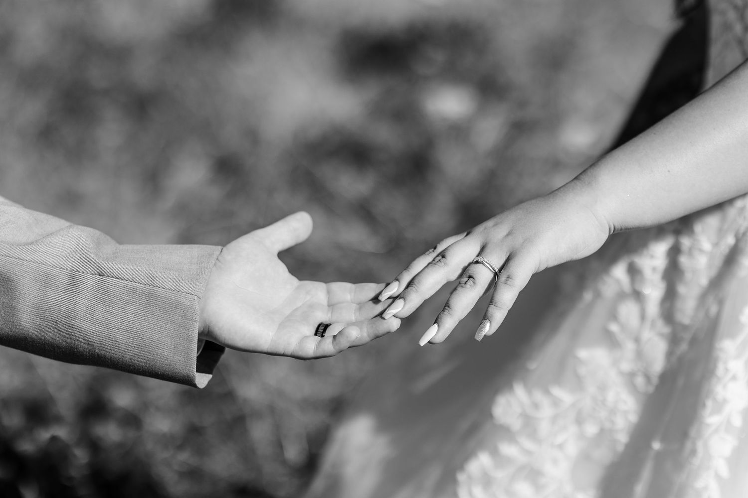 A black and white close-up photograph showing two hands reaching towards each other with a wedding ring visible.