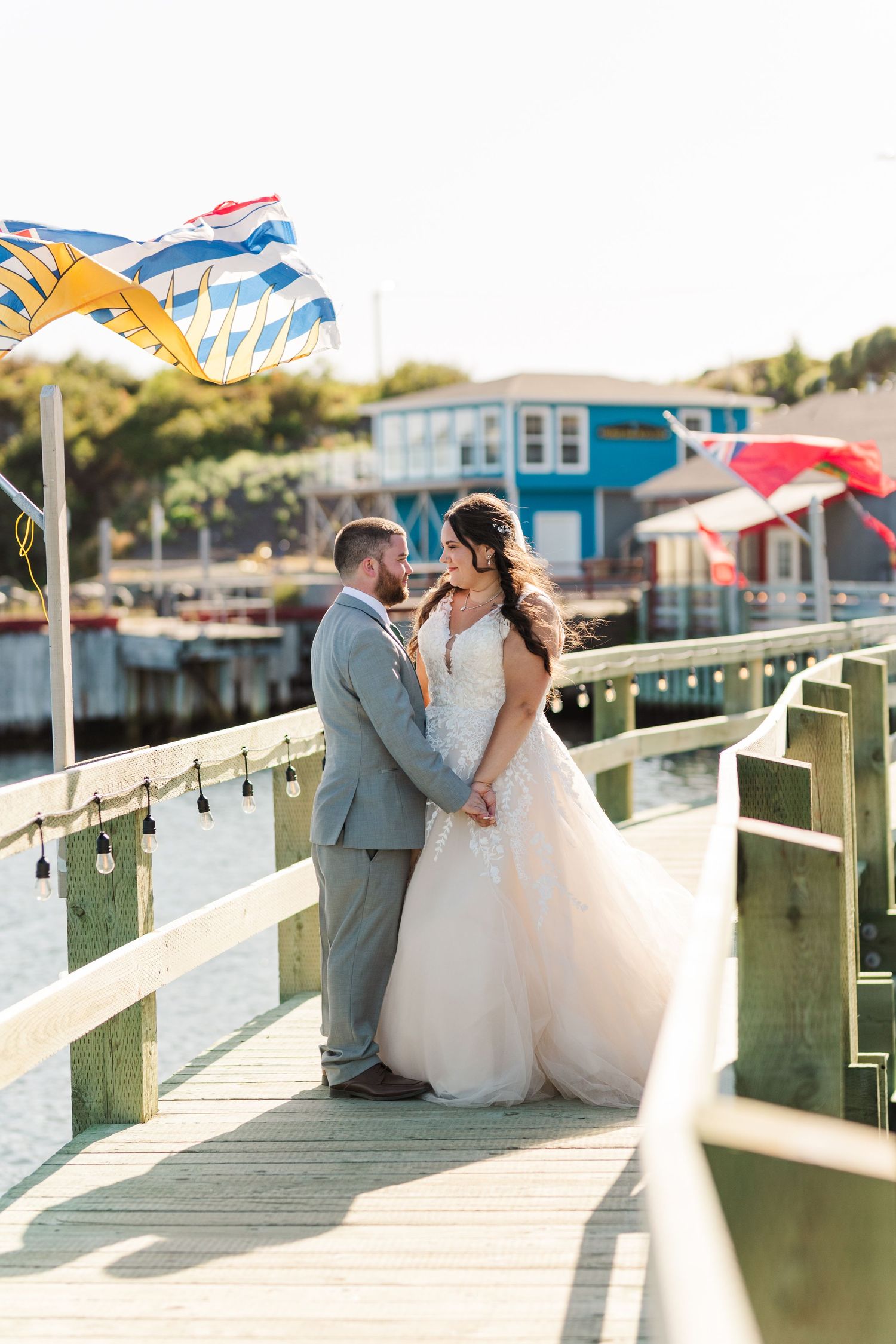 A couple shares a romantic moment on a waterfront dock with boats and a blue building in the background.