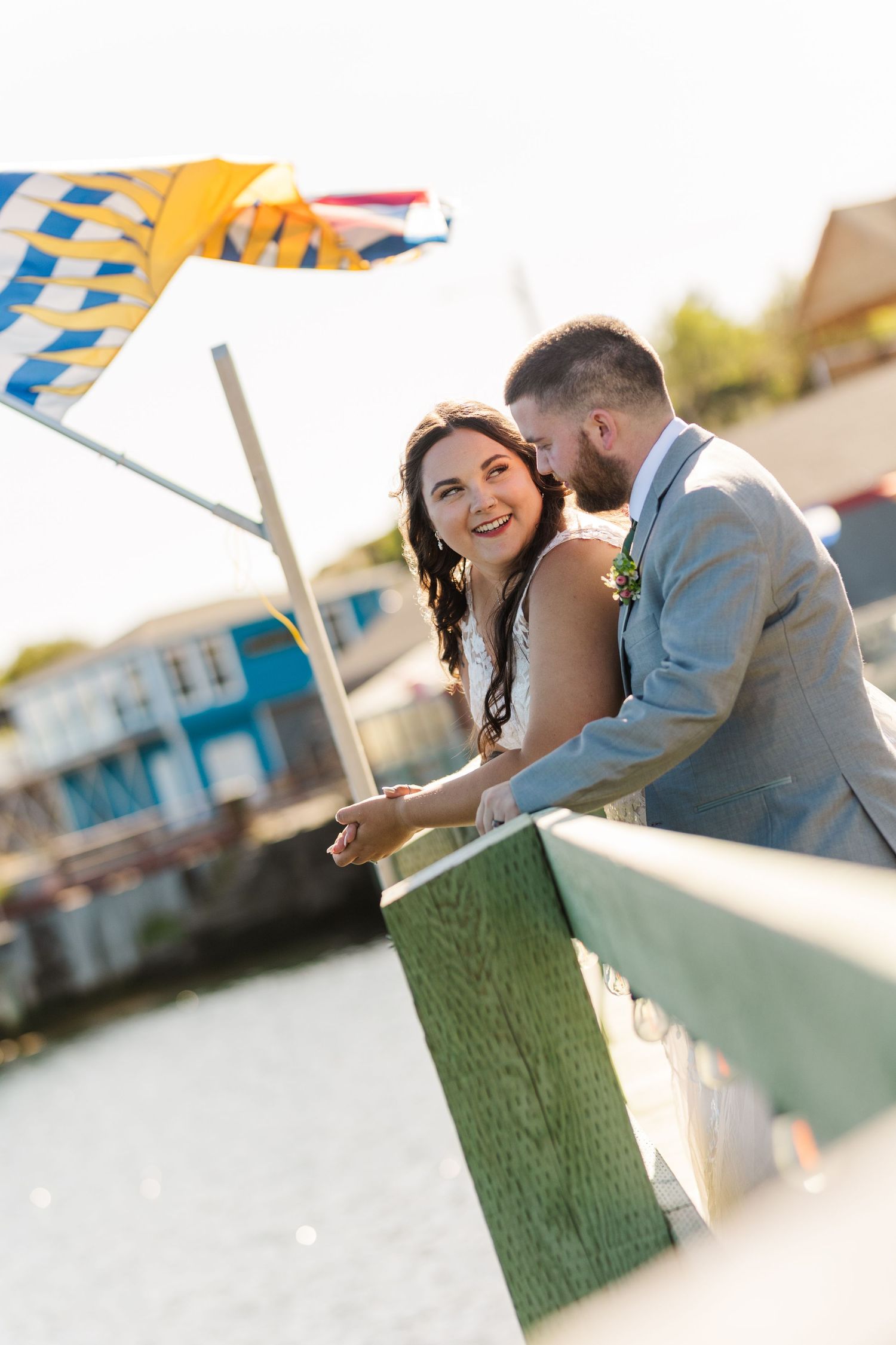 A couple in wedding attire share a romantic moment while leaning on a railing near a waterfront with boats in the background.