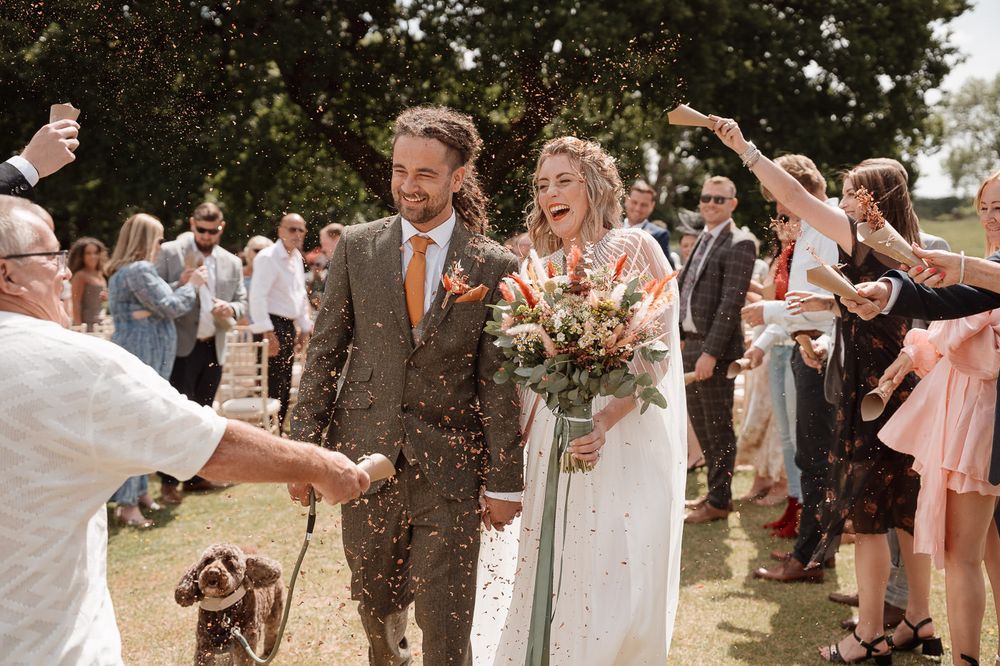 Wedding couple and guests celebrate outdoors with joyful gestures and confetti throwing on sunny summer day.