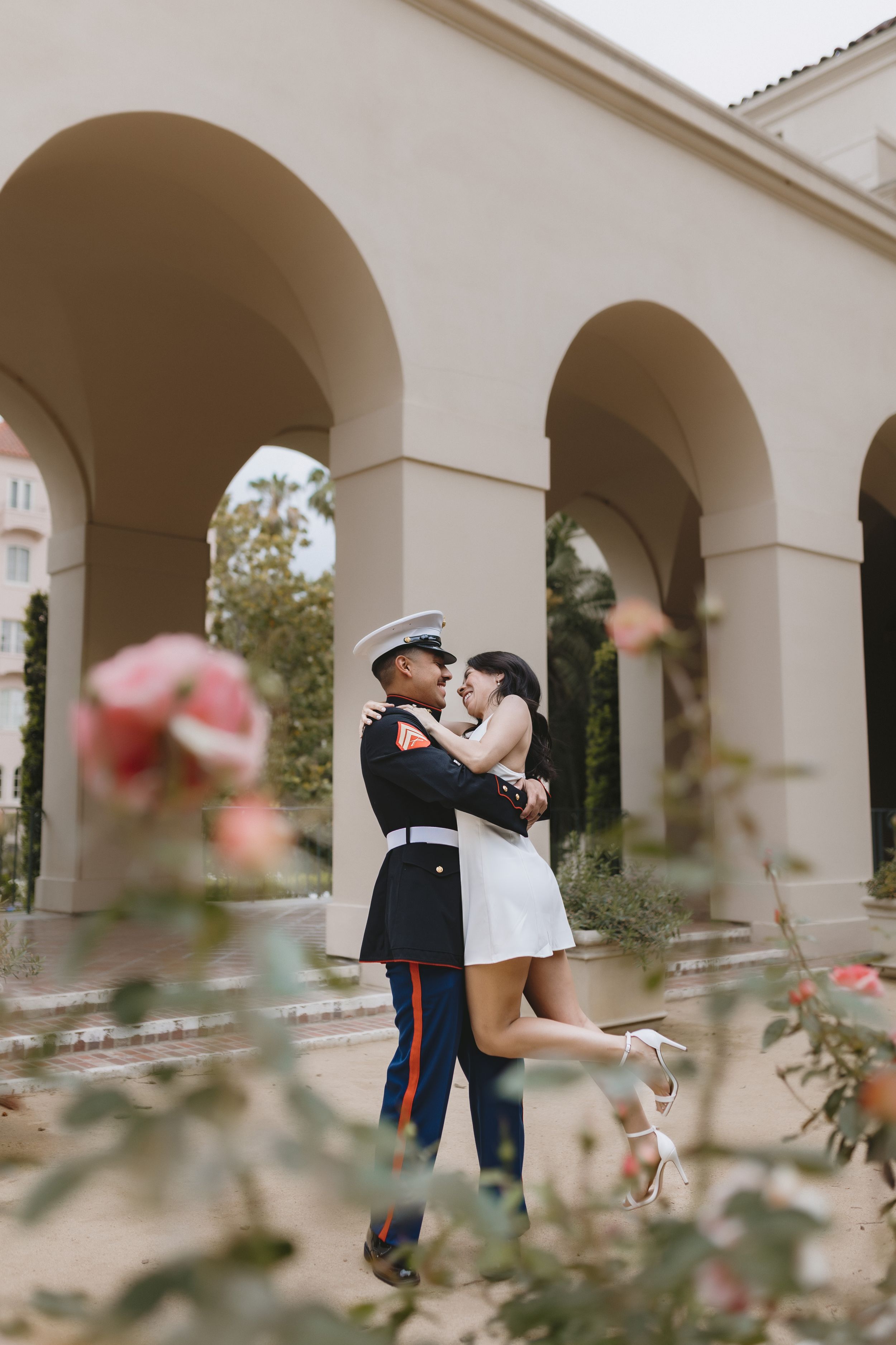 A romantic embrace between a couple under white arches, with pink roses in the foreground and Mediterranean architecture.