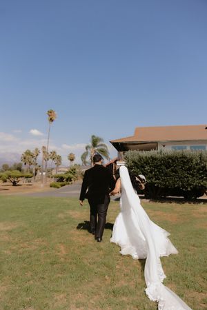 A couple in formal attire walks across a grassy field with palm trees and desert landscape in the background.
