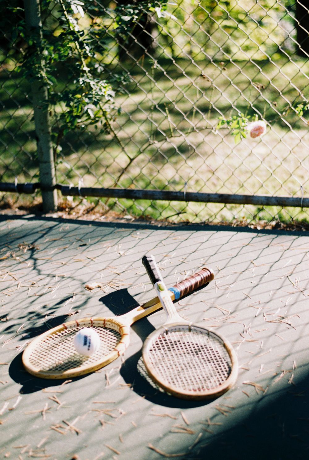 Four wooden tennis rackets cast shadows while arranged on a concrete surface in sunlight through a wire fence.