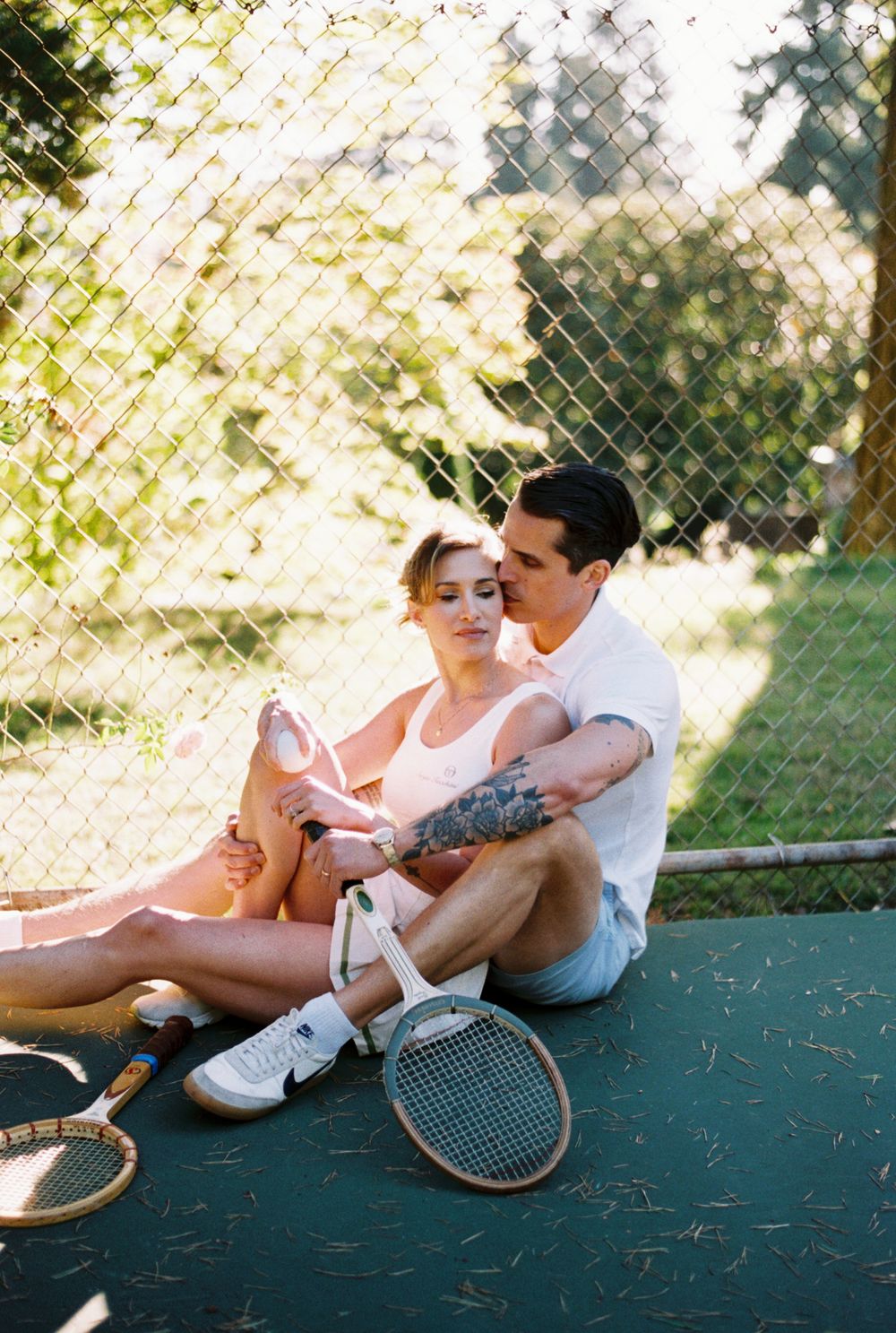 Romantic couple relaxes together with vintage tennis rackets on outdoor tennis court in sunlit setting.