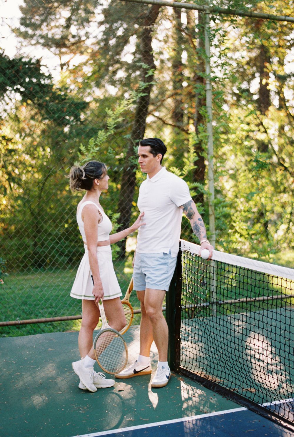 Two people in white tennis outfits share a playful moment on a blue tennis court surrounded by trees.