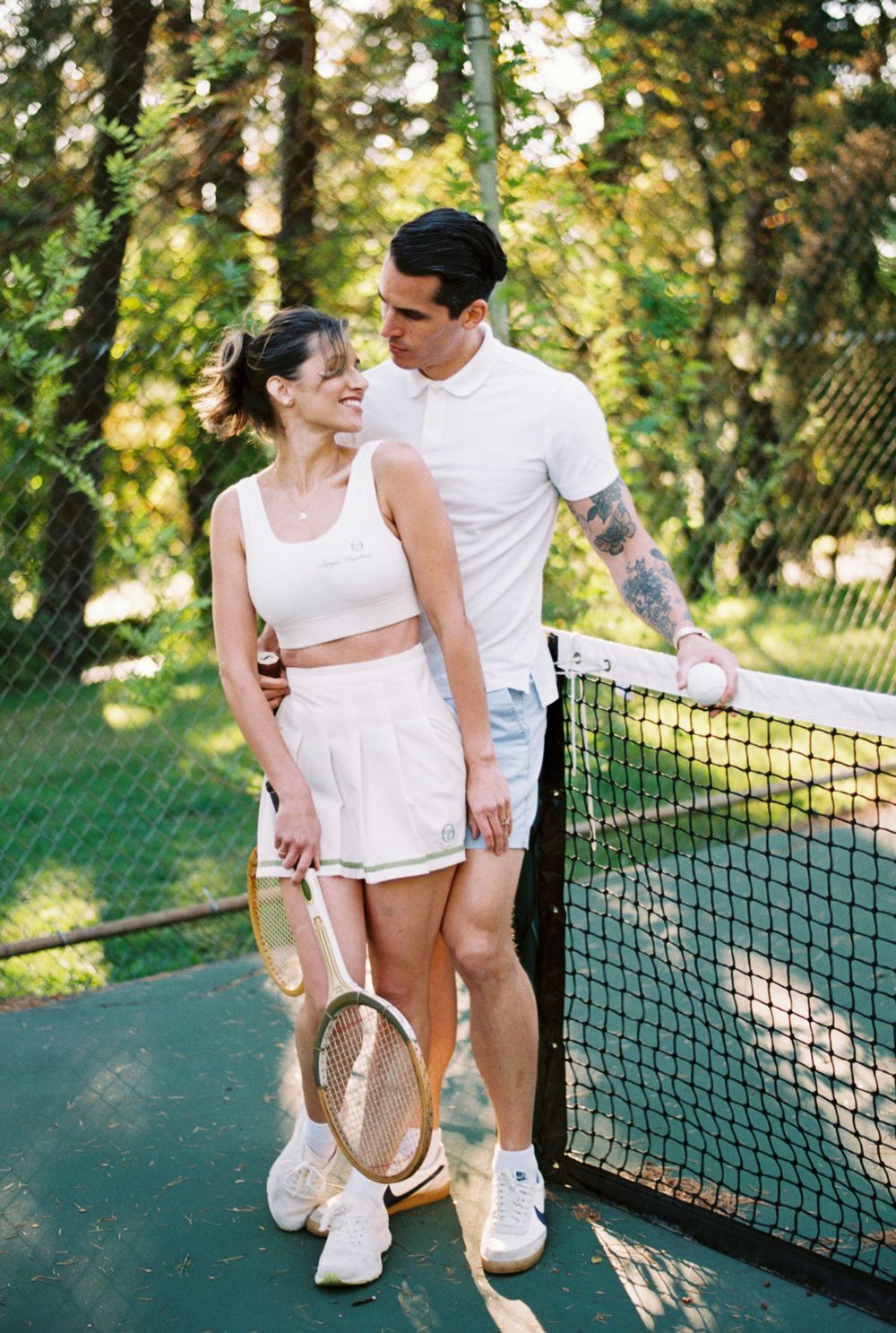 A couple shares an intimate moment on a tennis court while wearing matching white athletic outfits on a sunny day.