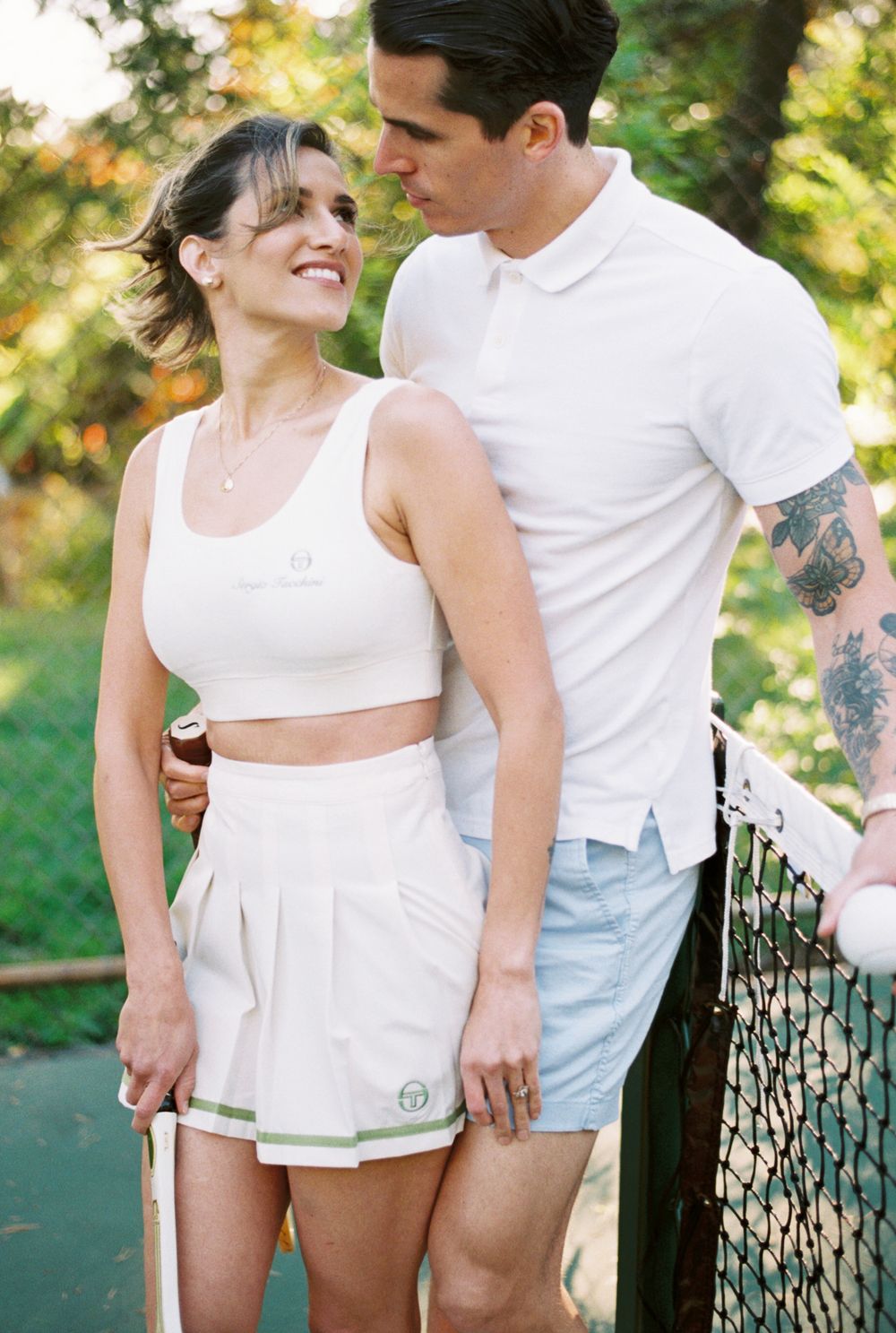 Couple in matching white outfits share intimate moments by tennis net in sunny outdoor setting.