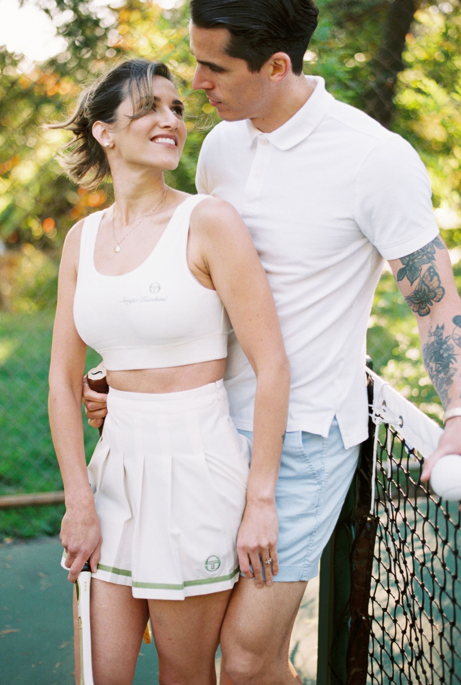 Couple in matching white outfits share intimate moments by tennis net in sunny outdoor setting.
