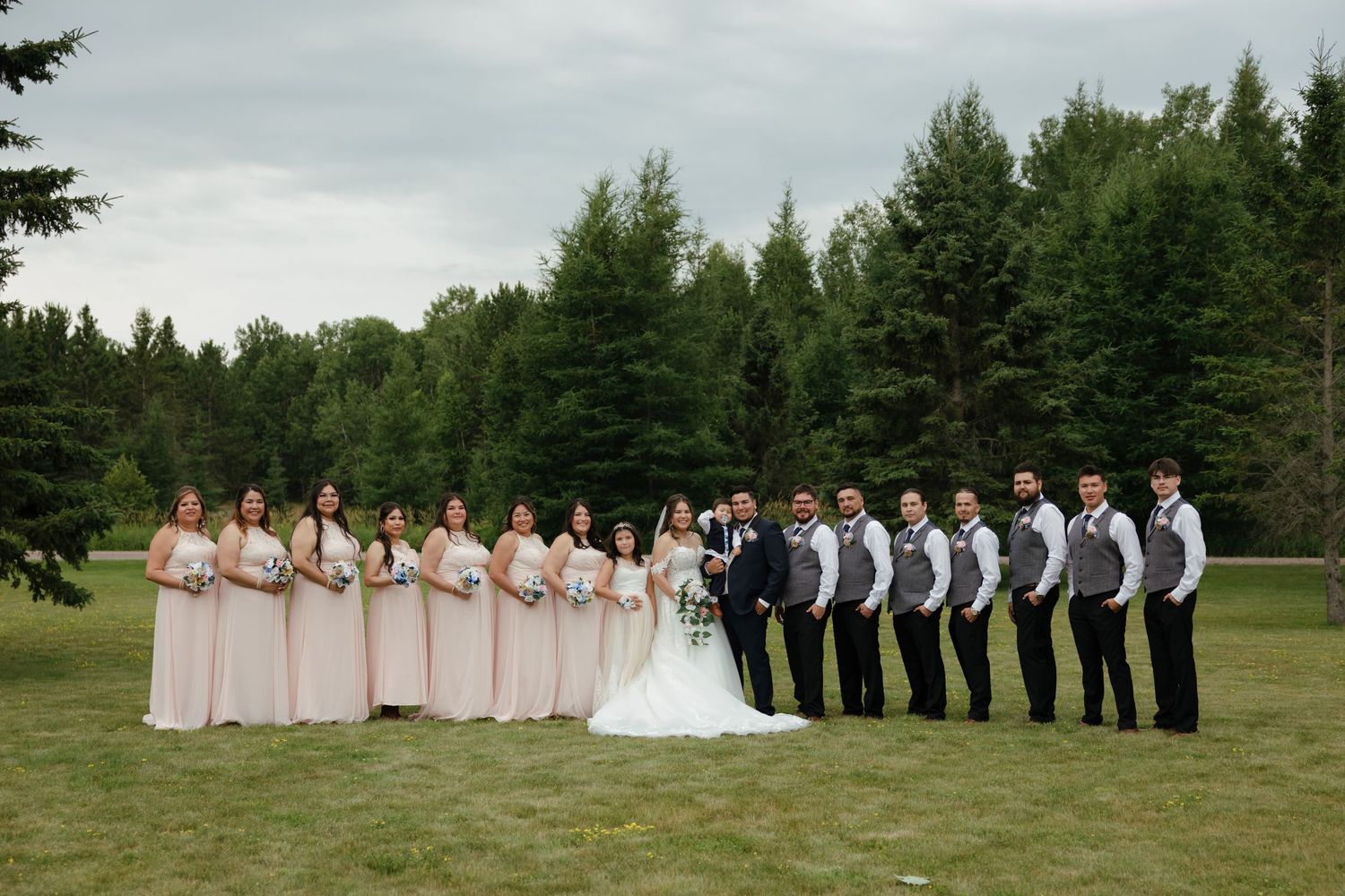 A wedding party stands outdoors in a grassy field surrounded by evergreen trees on a cloudy day.