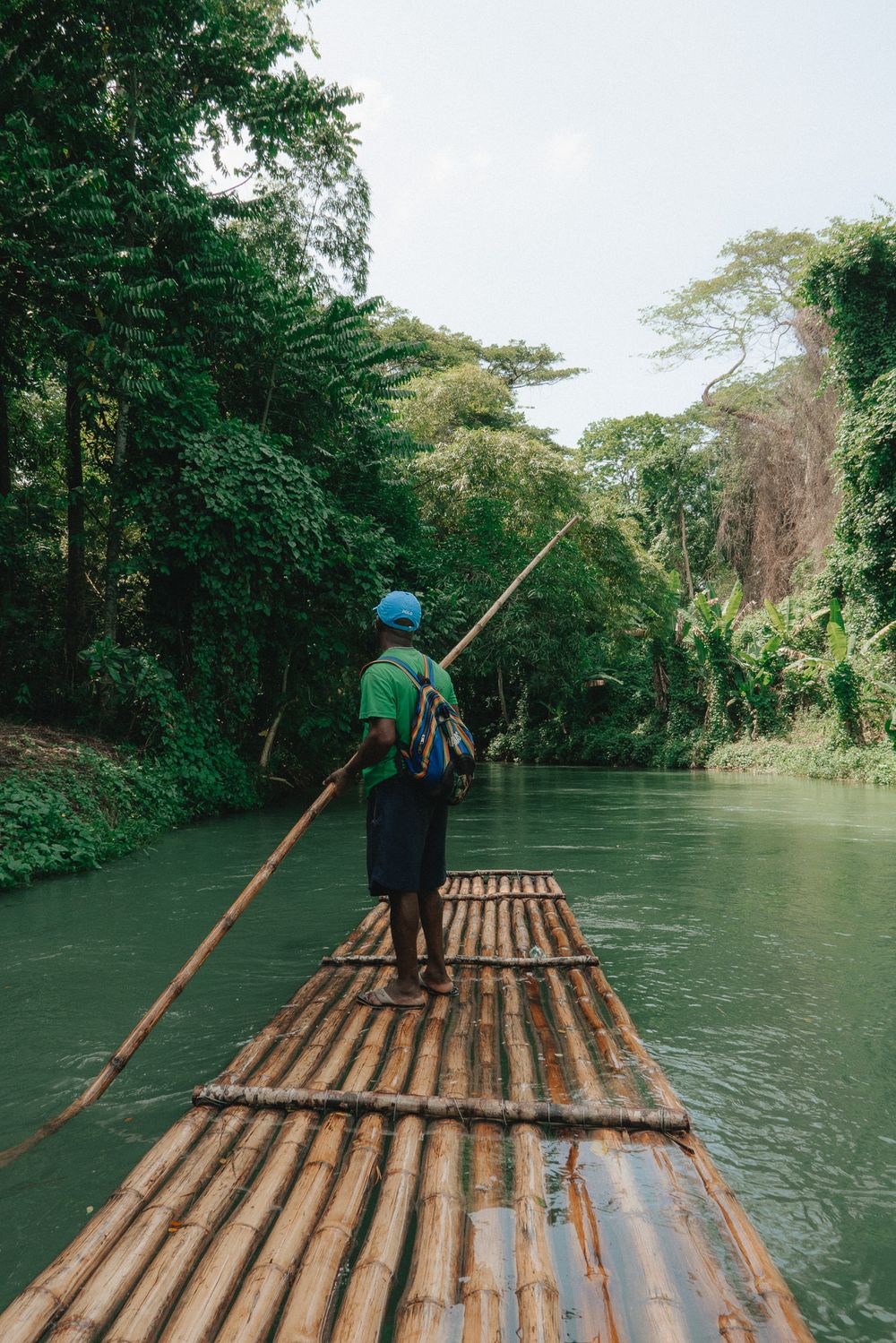 Person guides a traditional bamboo raft down a peaceful river surrounded by lush green jungle in Jamaica.