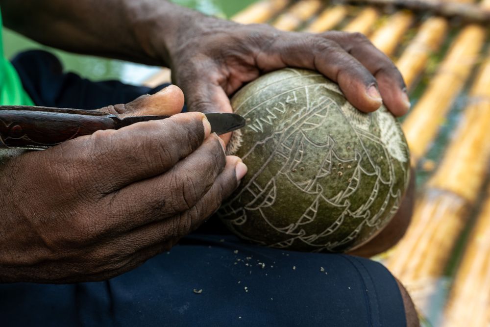 Hands delicately carving intricate patterns into a green coconut shell using traditional tools and techniques.