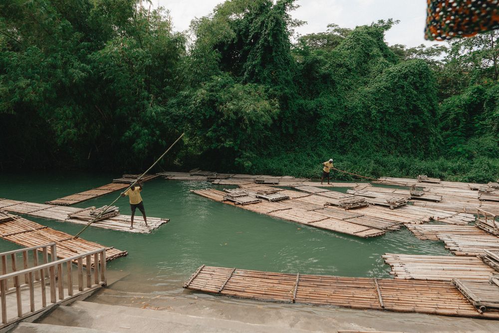 Bamboo rafts float on a green river surrounded by lush vegetation and rocks during a jungle excursion.