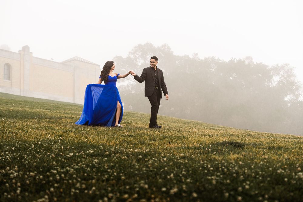 Couple in blue gown and black suit holding hands on a misty green field with trees and building backdrop.